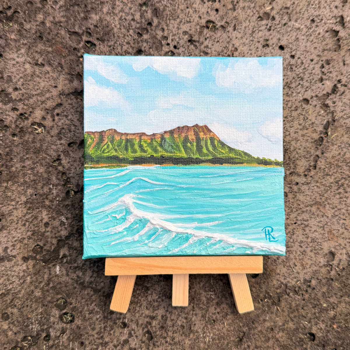 A small painted canvas showing a coastal landscape with turquoise water, a green and brown cliff, and a pale blue sky, displayed on a tiny wooden easel against a textured brown concrete surface.