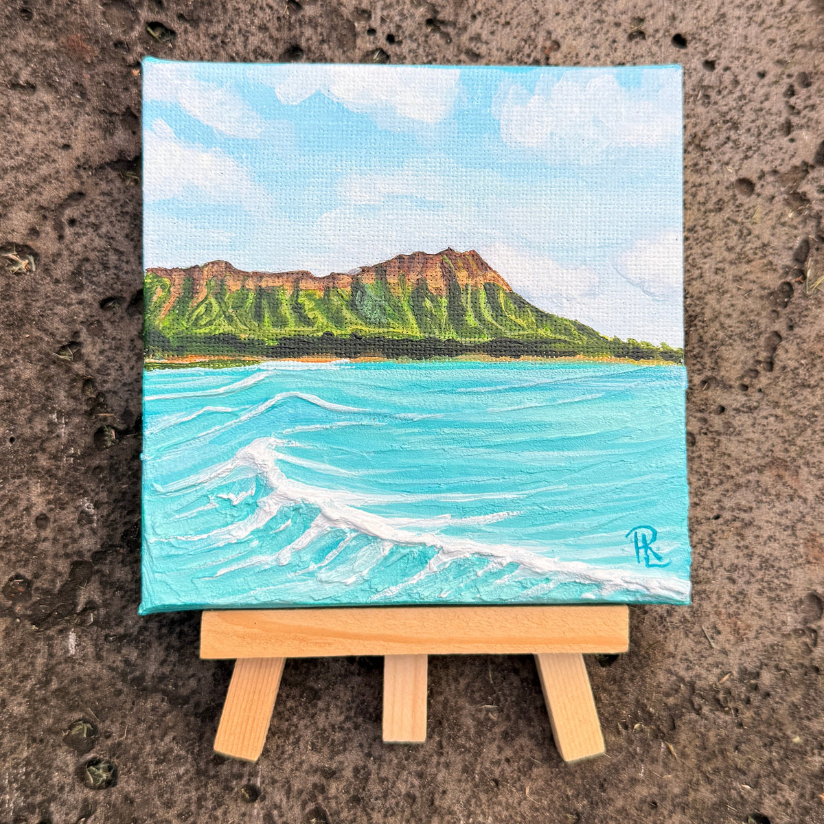 A small painted canvas showing a coastal landscape with turquoise water, diamond head, and a pale blue sky, displayed on a tiny wooden easel against a textured brown concrete surface.