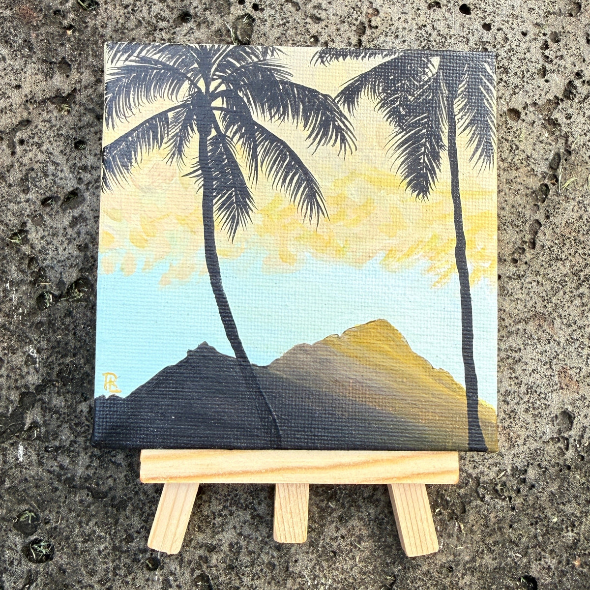 A small painted canvas showing palm trees, mountains, and a yellow sky, displayed on a miniature wooden easel against a textured concrete surface.