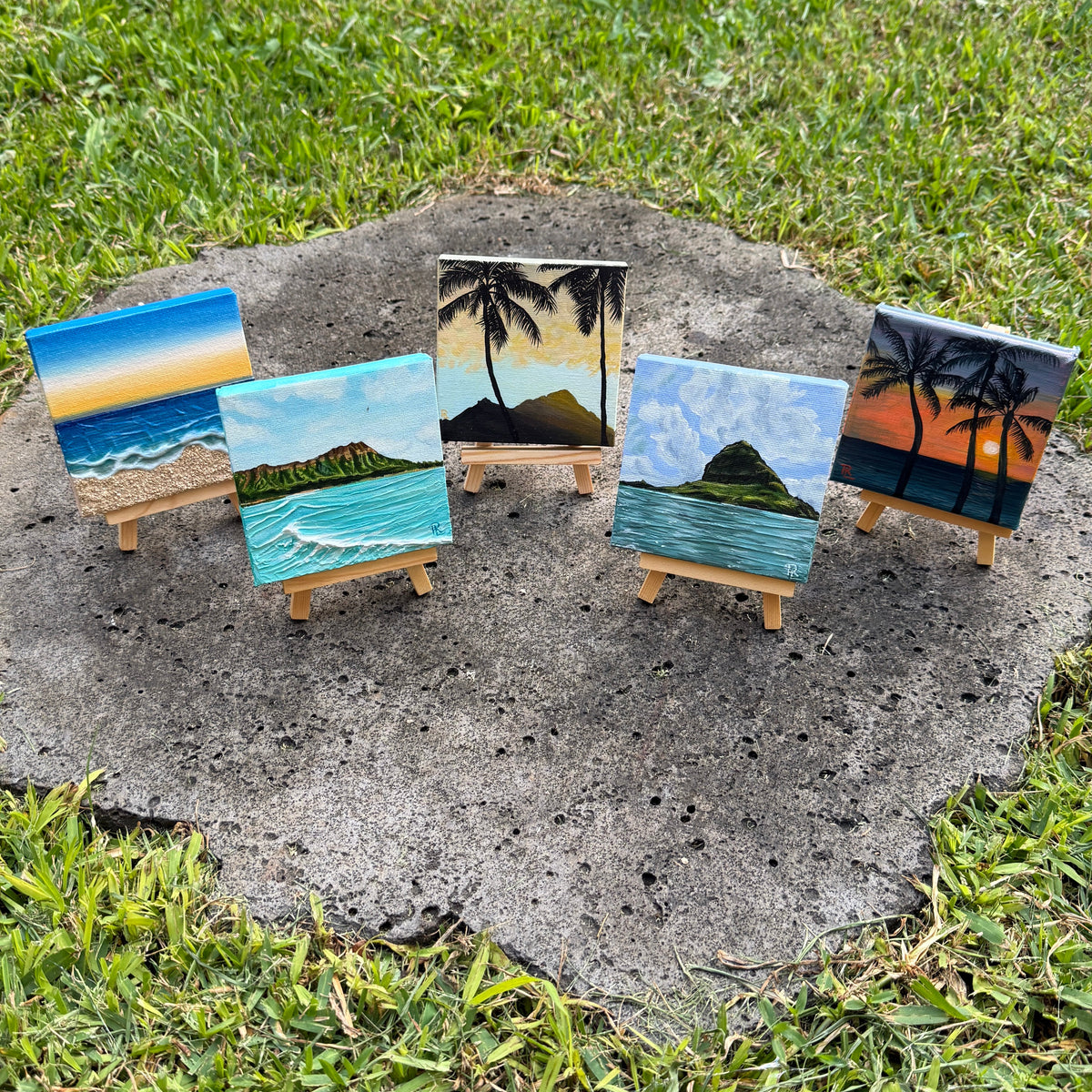 Five small hand-painted canvas prints of tropical beach and island scenes, each displayed on a miniature wooden easel outdoors