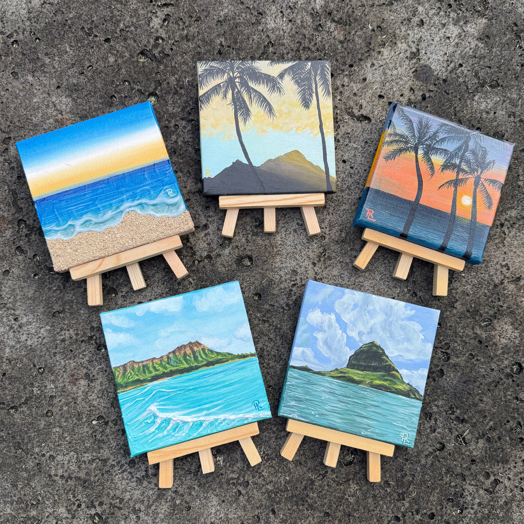 Five small hand-painted canvas art prints on wooden easels, arranged on a dark textured stone surface with grass visible in the corners. The canvases depict tropical beach scenes with blue ocean waves, sandy shores, cliffs, and palm trees.