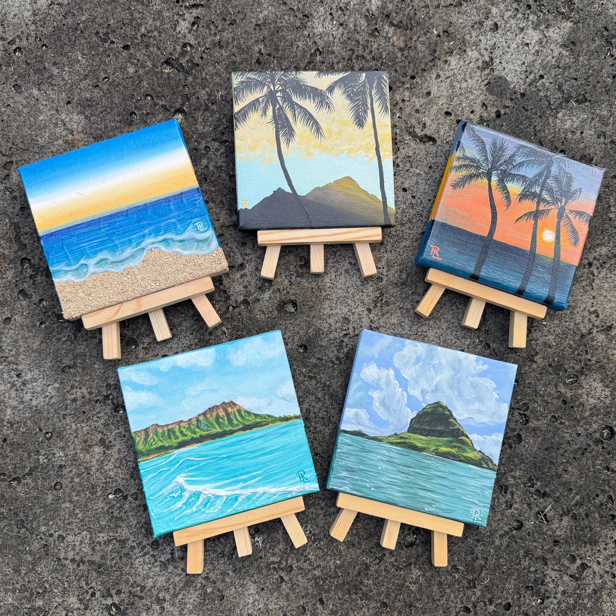 Five small hand-painted canvas art prints on wooden easels, arranged on a dark textured stone surface with grass visible in the corners. The canvases depict tropical beach scenes with blue ocean waves, sandy shores, cliffs, and palm trees.