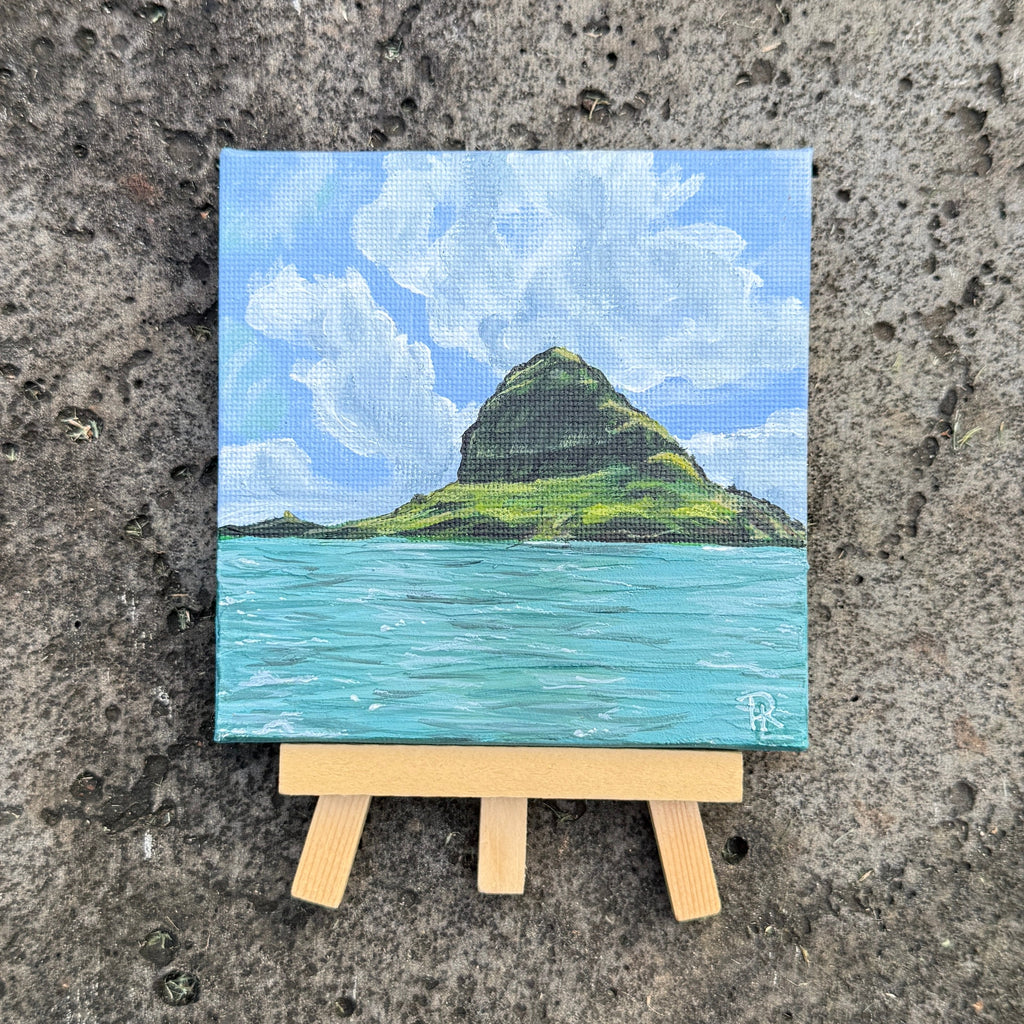 A small square canvas painting of a green island rising from turquoise water under a blue sky with white clouds, displayed on a miniature wooden easel against a textured concrete background.