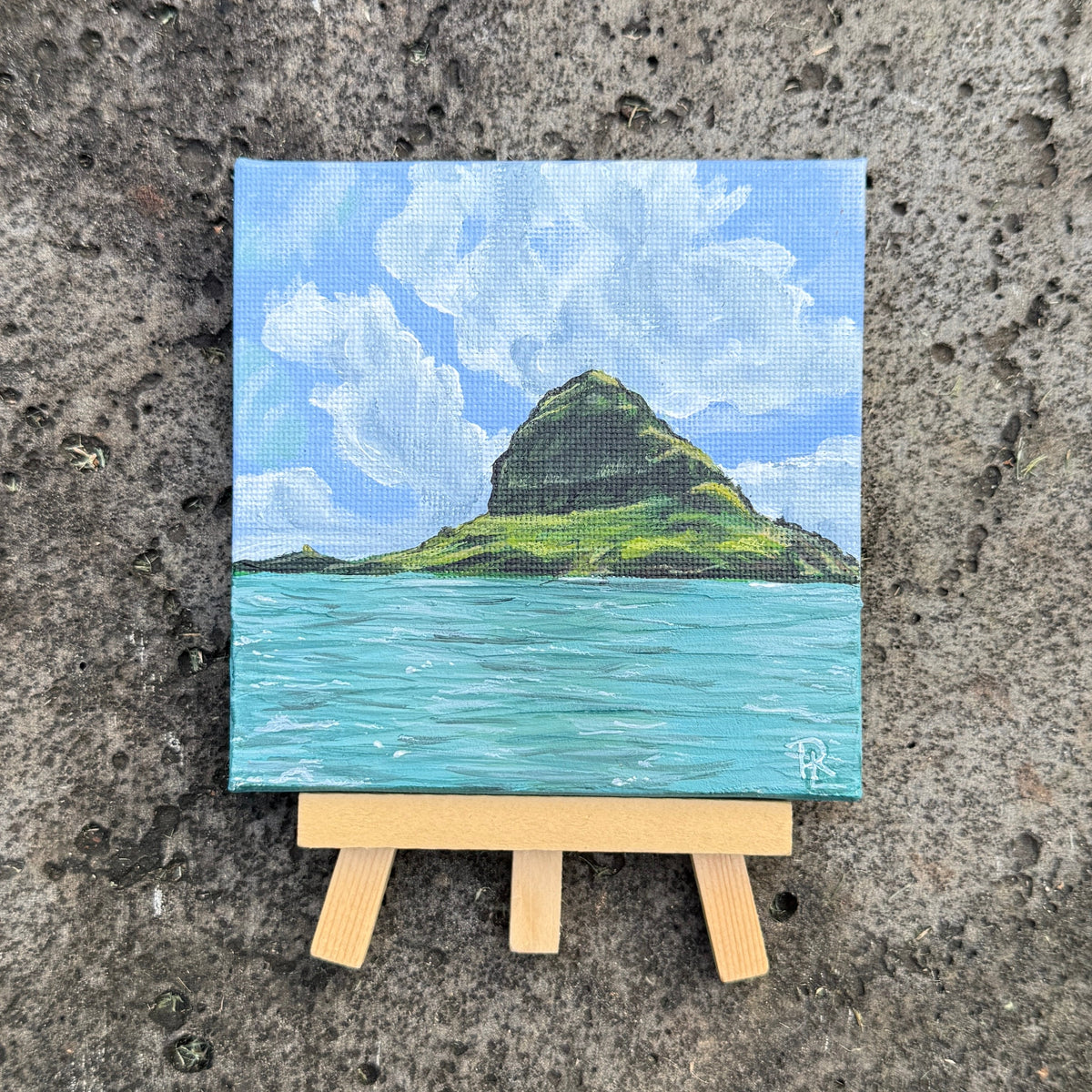 A small square canvas painting of a green island rising from turquoise water under a blue sky with white clouds, displayed on a miniature wooden easel against a textured concrete background.