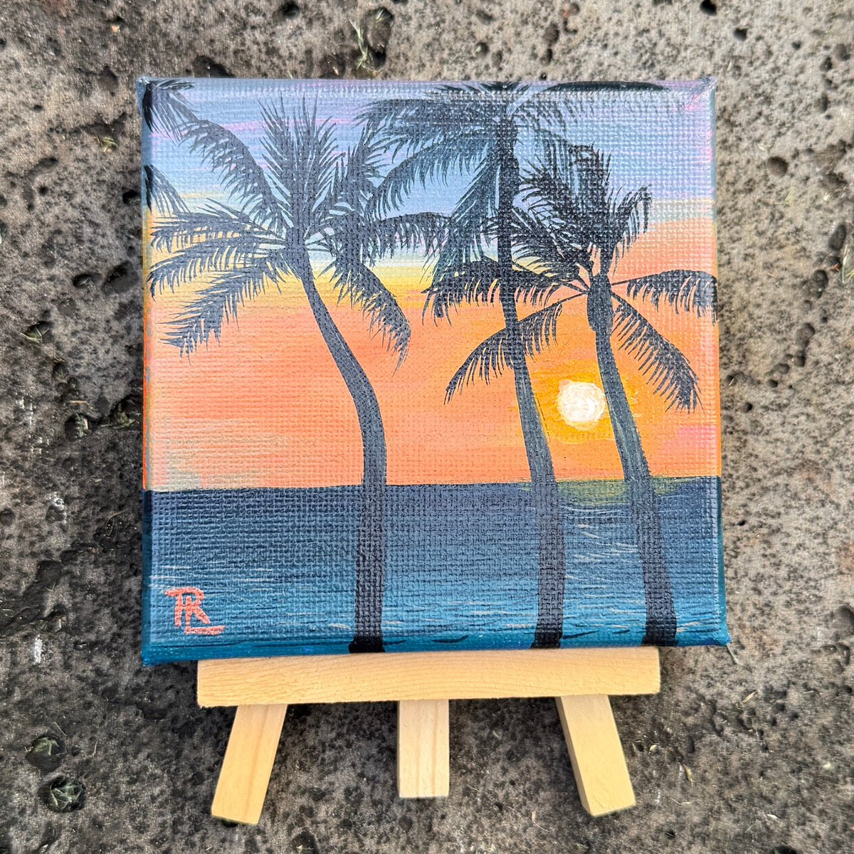 A small square canvas painting of a tropical sunset with palm trees over blue water, displayed on a miniature wooden easel against a textured concrete surface.
